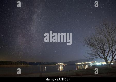 Milchstraße über einen blattlosen Baum am Ufer des Lake Jindabyne Jindabyne mit der Stadt im Hintergrund Stockfoto