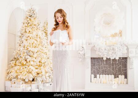 Schöne junge Frau in Silber Kleid öffnet Geschenk gegen den Hintergrund des Neuen Jahres Baum und Kamin, weißer Farbe. Traum Weihnachtsgeschenk Konzept Stockfoto