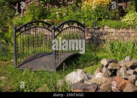 Alte schwarze schmiedeeiserne Metallbrücke und erhöhte Steingrenze mit gelben Rudbeckias, Hosta Pflanzen und lila Echinacea purpurea - Kegelblumen Stockfoto