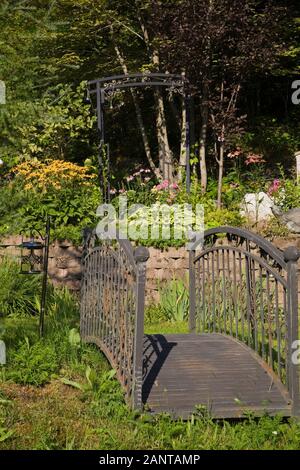 Alte schwarze schmiedeeiserne Metallbrücke und erhöhte Steingrenze mit gelben Rudbeckias, Hosta Pflanzen und lila Echinacea purpurea - Kegelblumen Stockfoto