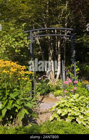 Flagstone Weg durch schwarzen schmiedeeisernen Metall Gartenlaube umgeben von gelben Rudbeckien - Coneflowers, Hosta Pflanzen im Vorgarten Land Garten im Sommer Stockfoto