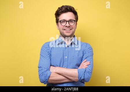 Hübscher junger Mann im blauen Hemd mit gefalteten Händen lächelnd, selbstbewusst gegen gelben Hintergrund Stockfoto