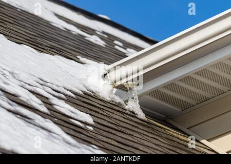 Schmelzender Schnee auf dem Dach des Hauses hat Eis auf Schindeln und Eiszapfen gebildet, die an der Dachrinne hängen Stockfoto