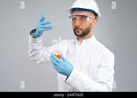 Vorderansicht des jungen Chemikers in Brille und Helm mit orangefarbener Flüssigkeit und Denken. Wissenschaftler in Handschuhen und weißem Labormantel experimentieren im Labor. Konzept der Chemie. Stockfoto