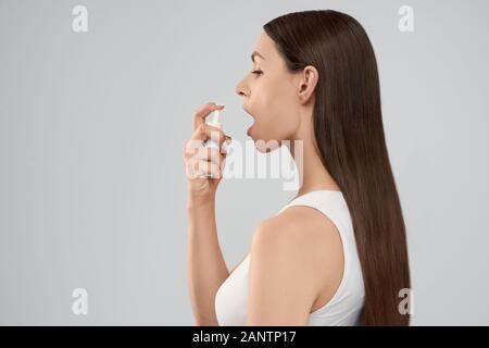 Seitenansicht der jungen hübschen brünetten Frau mit langem Haar mit Kehlspray. Isolierte Ernte von Frauen im Hemd, das sich posiert und weiße Flasche mit Medizin hält. Konzept der Medizin, Behandlung. Stockfoto