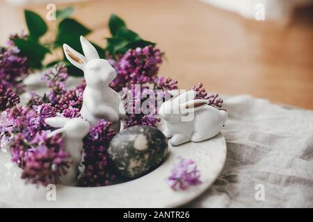 Frohe Ostern. Stilvolle Ostern weiße Häschen, moderne Osterei auf vintage Platte, und lila Blumen auf ländlichen Struktur. Natürlich gefärbte Ostereier und Feder Stockfoto