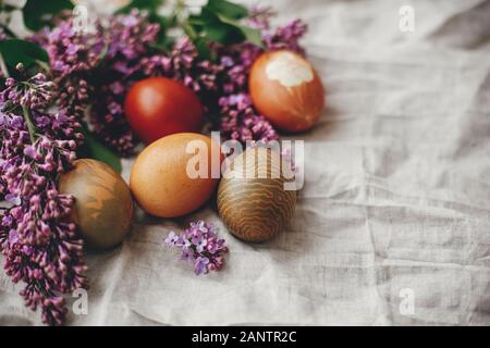 Moderne und farbenfrohe Ostereier auf rustikalen Leinen Stoff und Lila lila Blüten. Ländliche noch leben. Frohe Ostern. Platz für Text. Natürlich gefärbte Ostereier. Stockfoto