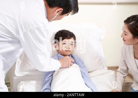 Fünf Jahre alten asiatischen Kind im Bett liegt im Krankenhaus von der Mutter begleitet und durch Kinderarzt mit Stethoskop untersucht Stockfoto