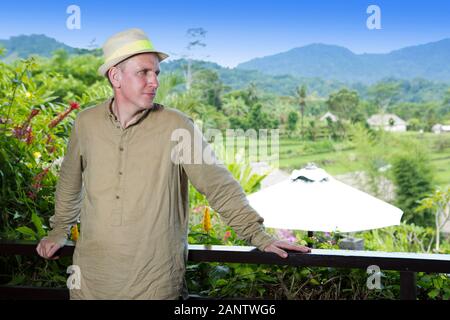 Reifer Mann im Sommer Kleidung und einen Sonnenhut auf der Veranda schaut auf die Tropen und Berge Stockfoto