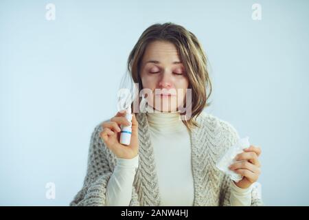 Kranke moderne 40 Jahre alte Frau in Rollkragen Pullover und Strickjacke mit Nasenspray auf Winter hellblauen Hintergrund Serviette. Stockfoto