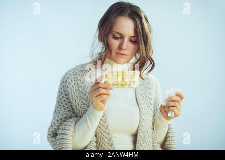 Kranke moderne Frau in Rollkragen Pullover und Strickjacke mit Serviette von Lesekompetenz auf Blister mit Pillen auf Winter hellblauen Hintergrund isoliert. Stockfoto