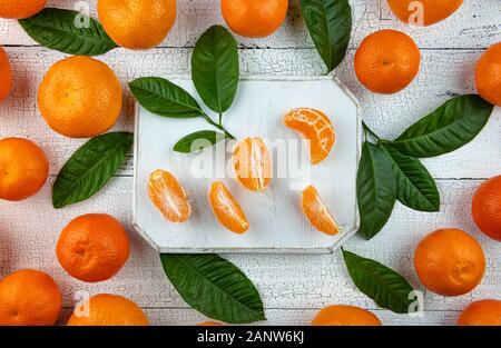 Tangerine Segmente auf Quadrat Weiß Schneidebrett von ganzen orange Tangerinen und Blätter auf alten Holztisch umgeben. Früchte und Blätter haben natürliche Stockfoto