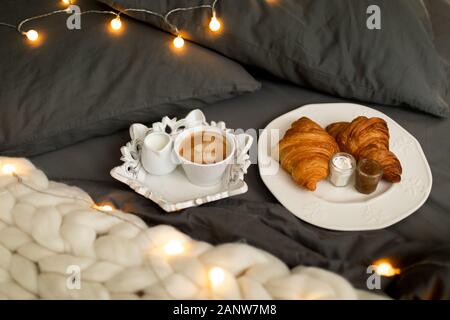 Frische Croissants mit Marmeladen und Americano mit Milch auf gestrickter weißer Wolldecke und leuchtenden Girlanden. Gemütlicher Wintermorgen zu Hause. Skandinavisches Schlafzimmer. Stockfoto