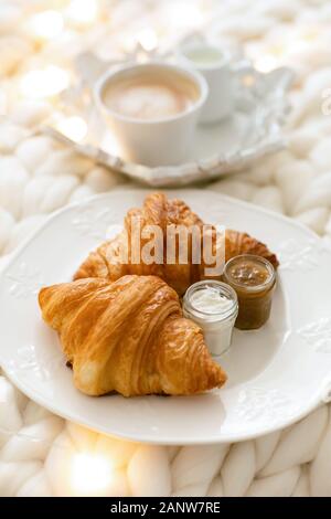 Frische Croissants mit Marmeladen und Americano mit Milch auf gestrickter weißer Wolldecke und leuchtenden Girlanden. Gemütlicher Wintermorgen zu Hause. Skandinavisches Schlafzimmer. Stockfoto