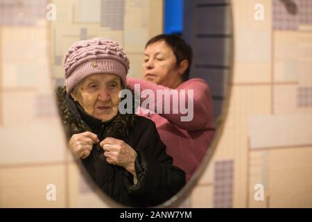 Reflexion im Spiegel, eine Frau im Mantel ihrer alten Mutter hilft. Stockfoto