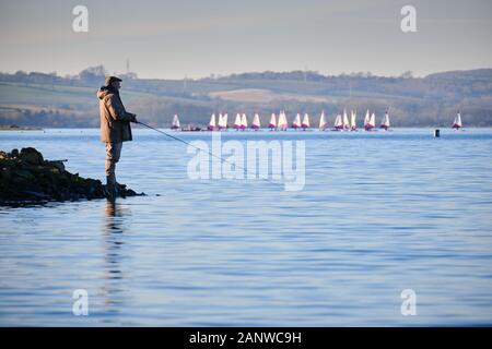 Angler-Fischer Sportfischen am Ufer des Rutland-Wassers ein anglianischer Wasserreservoir mit einer Flottille kleiner Segelboote im Hintergrund Stockfoto