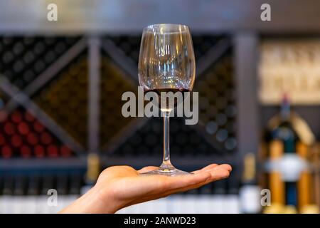 Ein Glas Rotwein an Hand in selektiven Fokus Nahaufnahme gegen Wein Flaschen auf X cube Lagerregale, Tasting Room interior Hintergrund verschwommen Stockfoto