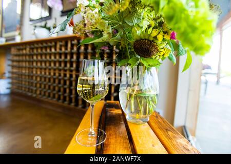 Ein gefülltes Glas Weißwein auf einen hölzernen Tisch neben einem Blumen Glas Vase selektiven Fokus, Wein Flaschenregale im Hintergrund, Weinproben Stockfoto