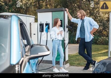 Reisen. Junges Paar reisen mit elektrischen Auto halt an der Ladestation sprechen surfen Smartphone betrachten einander glücklich, während sie darauf warten, Stockfoto