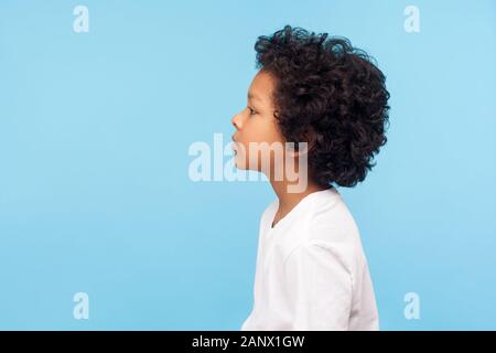 Profil von Netter kleiner Junge mit stilvollen lockiges Haar in T-Shirt auf der Suche mit schweren aufmerksames Gesicht zu Seite, ruhiger und nachdenklicher Ausdruck. indoor Stud Stockfoto