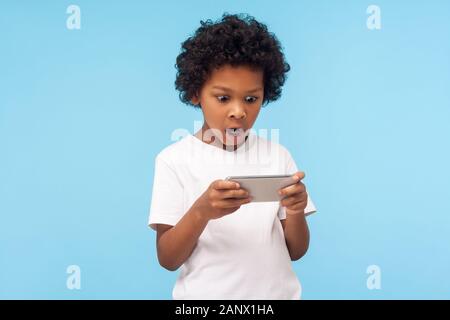 Überrascht, verblüfft Lustige kleine Junge mit lockigem Haar mit Handy mit Schockierten Ausdruck, erstaunt Kid spielen video game auf Handy, Kinder Stockfoto