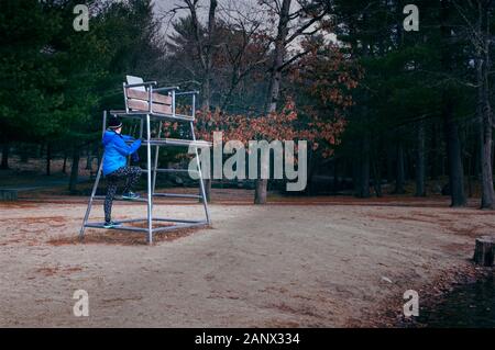 Eine Frau, die in der Fitness Kleidung Klettert eine Leiter zu einem Rettungsschwimmer Stuhl. Die Umgebung ist merklich Nebensaison (Herbst und Winter). Stockfoto