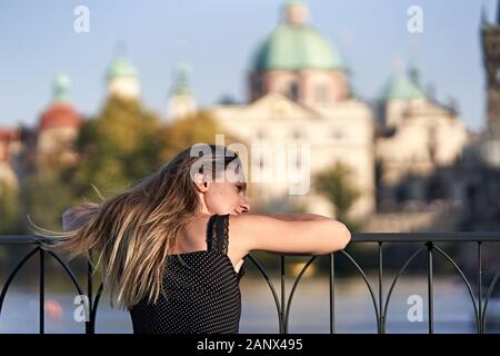 Junge attraktive Frau, die auf ihren Armen auf einem schwarzen Metallgeländer vor dem historischen alten Zentrum von Prag und dem Fluss ruht. Stockfoto