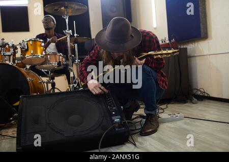 In voller Länge Porträt des zeitgenössischen Musiker einrichten Gitarre Verstärker beim Soundcheck während der Vorbereitung für Konzert oder Probe mit Band im Hintergrund, kopieren Raum Stockfoto