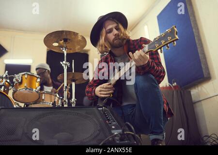 Low Angle Porträt des zeitgenössischen Langhaarige Musiker einrichten Gitarre beim Soundcheck während der Vorbereitung für Konzert oder Probe mit Band im Hintergrund, kopieren Raum Stockfoto