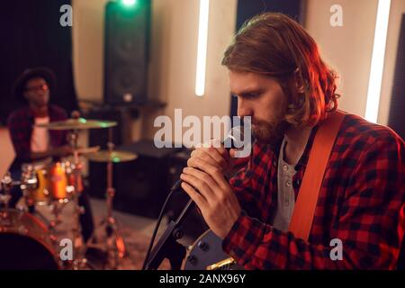 Seitenansicht Portrait von Stattlichen bärtiger Mann singen auf das Mikrofon während der Probe oder ein Konzert mit Musik Band in schwach beleuchteten Raum Studio, kopieren Stockfoto