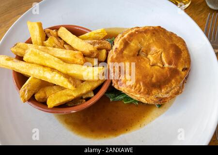 Typische deftig, lecker British Pub grub Mahlzeit: warmes Fleisch Torte in Soße mit einer Schüssel mit Golden gebratene chunky Chips auf ein weißes Porzellan Teller serviert Stockfoto