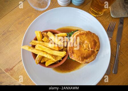 Typische deftig, lecker British Pub grub Mahlzeit: warmes Fleisch Torte in Soße mit einer Schüssel mit Golden gebratene chunky Chips auf ein weißes Porzellan Teller serviert Stockfoto