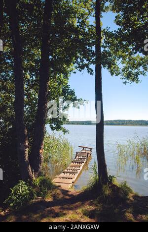 Die alten hölzernen Pier auf der See ist von Wasser, Bäumen und Schilf umgeben. Das Konzept der Entspannung und der Einheit mit der Natur. Stockfoto