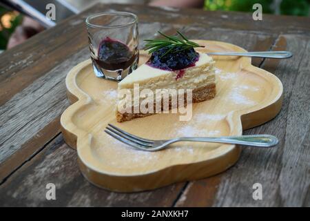 Käsekuchen Kreissegment mit blueberry Sirup mit Löffel und Gabel auf einem hölzernen Platte gekrönt, süße Speisen mit braunen Tisch im Hintergrund Stockfoto