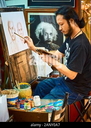 Bangkok, Thailand: Junger Mann sitzt an einem Markt der Malerei ein Portrait von ein paar; ein Bild von einem beobachten Einstein an der Wand hängen Stockfoto