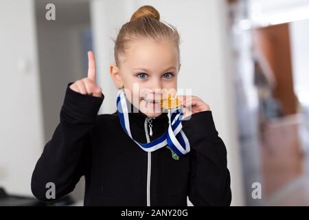 Porträt eines kleinen Mädchens - Gymnast in einen Trainingsanzug mit Medaillen am Hals, beißen die Medaille und Daumen hoch Stockfoto