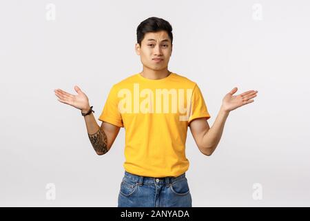 Skeptisch und Mühe unglücklich Junge arrogante asiatischer Mann mit Tätowierungen, tragen gelbe T-Shirt, breitete die Hände seitlich in Bestürzung und Verwirrung, Stockfoto