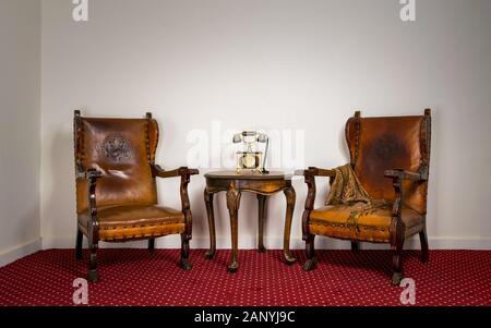 Zusammensetzung von zwei hölzernen Sessel, alten Telefon auf kleinen runden Tisch auf dem roten Teppich und weiße Wand gesetzt Stockfoto