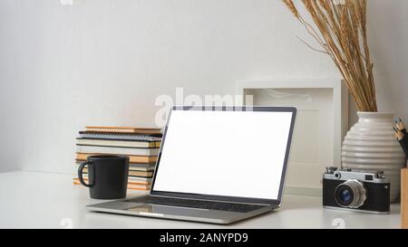 7/8 Schuß von Fotograf Arbeitsplatz mit leerer Bildschirm Laptop, Kamera, Rahmen, Büchern und Dekorationen auf weißem Schreibtisch mit weißen Wand Hintergrund Stockfoto