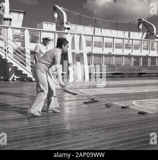 1960, historische, zwei Kreuzfahrtschiff Passagiere spielen ein Spiel der huffleboard', wo die Spieler lange Stöcke mit gebogenen Spitzen durch Gummi Festplatten über ein Gericht auf das Deck gemalt zu schieben. Es ist eine unterhaltsame Art und Weise für Passagiere Zeit draußen auf dem Meer Tage zu verbringen. Stockfoto