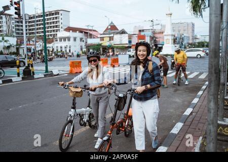 Zwei asiatische junge Frauen, die Helm und Taschen tragen, spazieren mit Klapprad Stockfoto