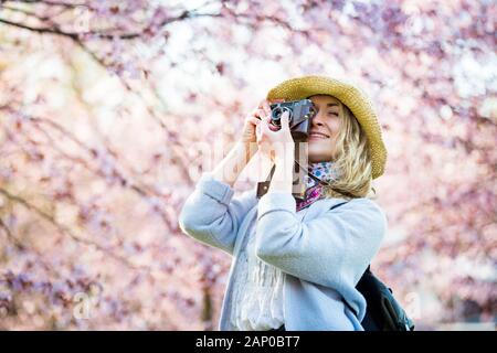 Porträt der schönen Frau in Strohhut Reise in einem schönen Park mit Kirschbäumen in Blüte, Fotos auf Retro-Kamera. Tourist mit Rucksack