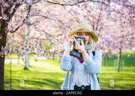 Porträt der schönen Frau in Strohhut Reise in einem schönen Park mit Kirschbäumen in Blüte, Fotos auf Retro-Kamera. Tourist mit Rucksack Stockfoto