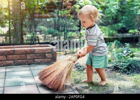 Cute adorable kaukasischen Kleinkind Junge spielt Holding Besen im Hinterhof in Garten im Freien. Kind little Helper in t-short Kurze Hose und Spaß haben Stockfoto
