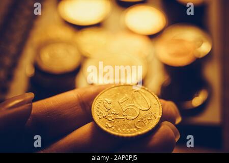 Goldene 50 Euro Cent Münze in der menschlichen Hand, Geld in weiblichen Fingern, Stapel von Münzen und Tisch im Hintergrund. Stockfoto