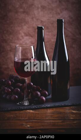 Glas Rotwein und zwei Flaschen mit Trauben auf einem Holztisch Stockfoto