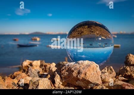 Tensphy Kristallkugel (Lensball) in einer Ruiene am Strand des Mar Menor (La Manga in Spanien) mit Schiffen, Booten, Steinen, Wasser, Sand und blauem Himmel. 25.12.19 | Verwendung weltweit Stockfoto