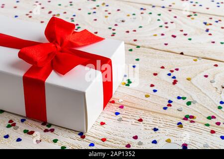 Weiß Geschenkbox mit roter Schleife auf einem weißen Holz strukturierte Oberfläche mit Herz Form Folie Konfetti. Gruß Valentine Karte. Romantik und Liebe Konzept. Stockfoto