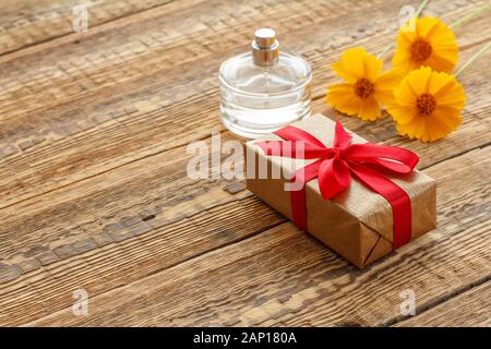 Geschenkbox mit roter Schleife und Flasche Parfüm auf Holzbrettern mit gelben Blüten gewickelt. Ansicht von oben. Stockfoto