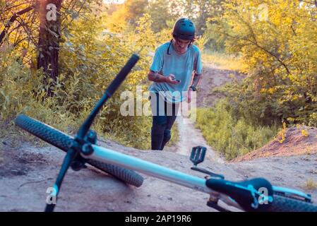 Der Radfahrer mit dem Smartphone in seinen Händen, ist zu Fuß. Auf dem Hintergrund liegt ein Fahrrad. Im Wald, im Freien. In warmen Farben Stockfoto